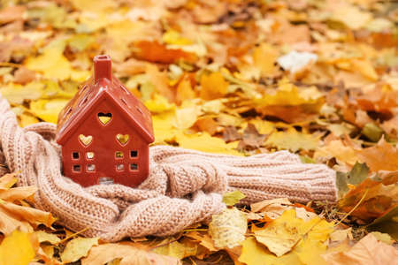 Figure of house and warm scarf on autumn leaves outdoors. Concept of heating seasonの写真素材
