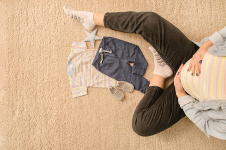 Pregnant woman with baby clothes sitting on floor at home