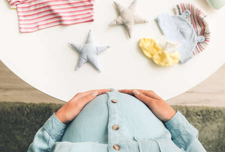Beautiful pregnant woman near table with baby clothes, top viewの写真素材