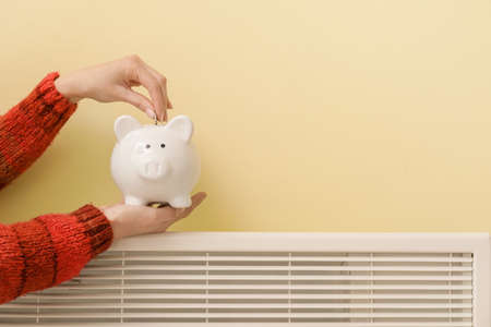 Woman putting money in piggy bank on radiator. Concept of heating seasonの写真素材