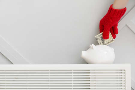 Woman putting money in piggy bank on radiator. Concept of heating seasonの写真素材