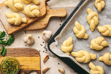 Baking sheet and board with uncooked garlic buns on light background