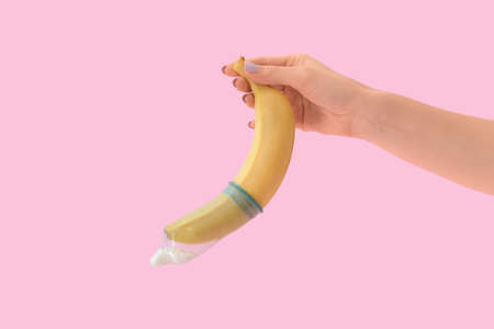 Female hand with banana in condom on color background. Erotic concept