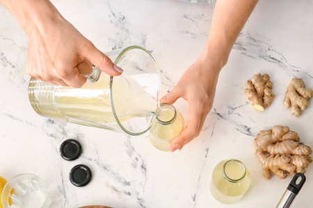 Woman pouring healthy ginger juice from jug into bottle on light background