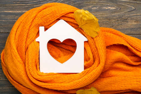 Figure of house, autumn leaves and warm scarf on wooden background. Concept of heating seasonの写真素材