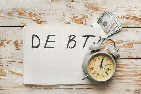 Alarm clock with money and notebook with word DEBT on white wooden backgroundの写真素材