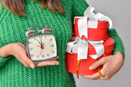 Young woman with alarm clock and Christmas gifts on gray background, closeup
