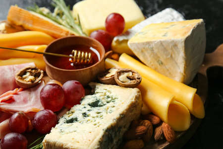 Board with tasty cheese, honey and snacks on dark background, closeup