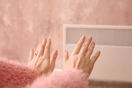 Young woman warming hands near electric heater at home. Concept of heating seasonの写真素材