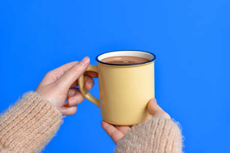 Hands of young woman with hot cocoa on color background
