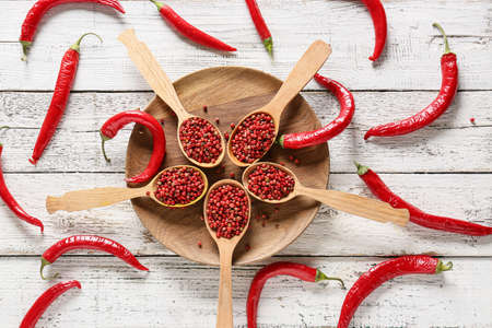 Spoons with red peppercorns on white wooden background