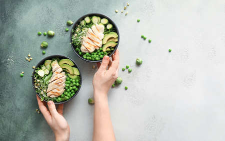 Female hands and bowls of tasty salad with fresh vegetables and chicken on light background