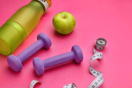 Dumbbells, measuring tape, bottle of water and apple on pink background