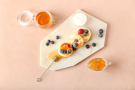 Board with tasty cottage cheese pancakes, berries and honey on color background
