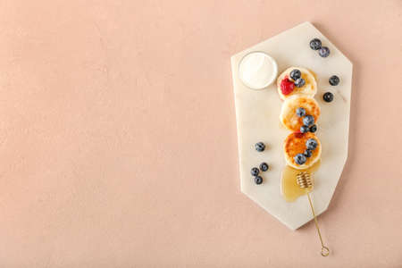 Board with tasty cottage cheese pancakes, berries and honey on color background