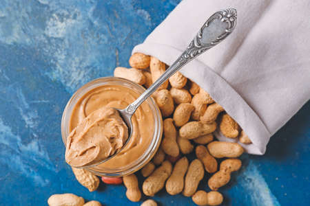 Jar of tasty peanut butter and sack with nuts on color background