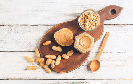 Jar with tasty peanut butter, bread and nuts on light wooden background