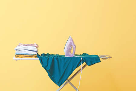 Board with electric iron and clean clothes on color background