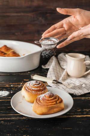 Woman sprinkling powdered sugar onto tasty cinnamon rolls on tableの写真素材