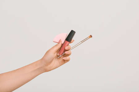 Female hand with stylish makeup sponge, brush and lipstick on light backgroundの写真素材