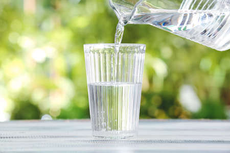 Pouring fresh water from jar into glass on table outdoors, closeup