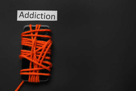Mobile phone tied with rope and word ADDICTION on dark background