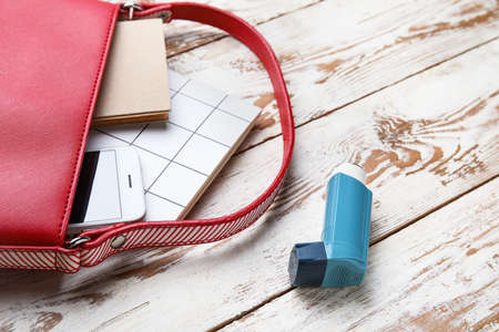 Stylish bag with inhaler and mobile phone on light wooden background