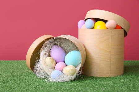 Boxes with beautiful Easter eggs on grass against color background