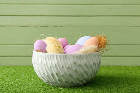 Bowl with beautiful Easter eggs on grass against color wooden background