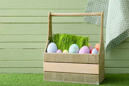 Basket with beautiful Easter eggs on grass against color wooden background