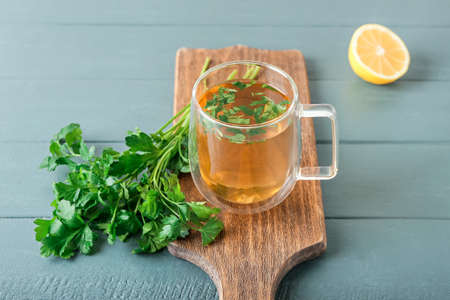 Board with cup of hot tea with parsley on color wooden background