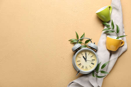 Composition with alarm clock, cups and green leaves on color background