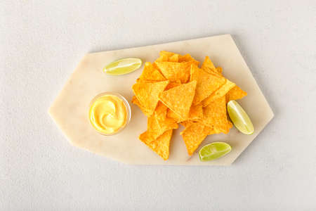 Board with tasty nachos, sauce and lime on light background