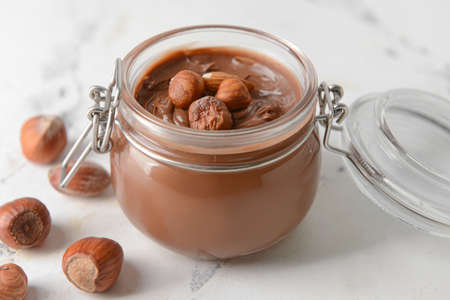 Jar with tasty chocolate paste and hazelnuts on light background