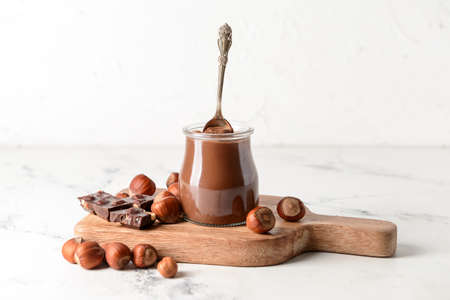 Jar with tasty chocolate paste and hazelnuts on light background