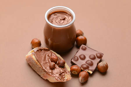 Jar and bread with tasty chocolate paste and hazelnuts on color background