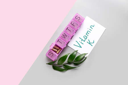 Paper with text VITAMIN K and organizer with pills on color background