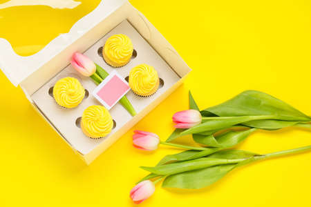 Box with tasty cupcakes and tulip flowers on color background