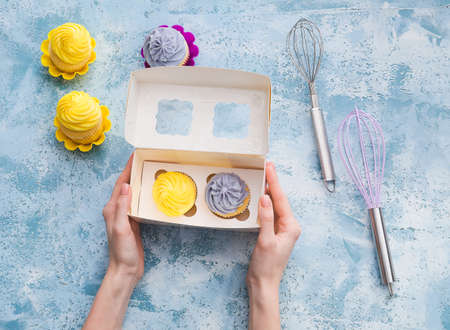 Female hands with tasty cupcakes in box on color background