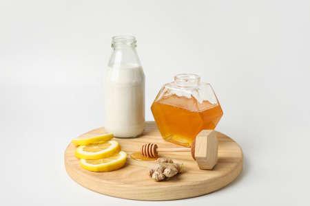 Jar with sweet honey, lemon slices, bottle of milk and ginger on wooden board against white...