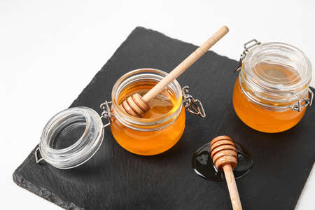 Glass jars with sweet honey and dippers on slate board against white background