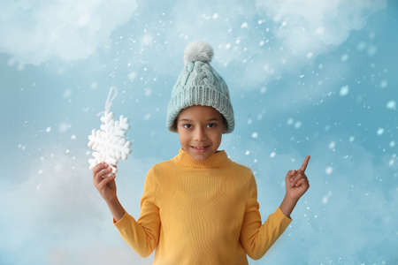 Cute little African-American boy in winter clothes and with snowflake showing something on color background with falling snowの写真素材