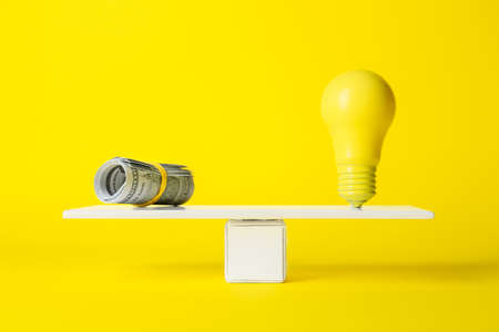 Dollar banknotes and light bulb on teeterboard against yellow background. Concept of balanceの写真素材