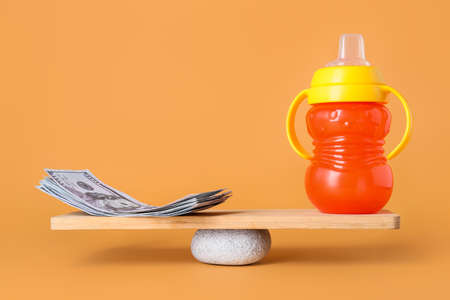 Dollar banknotes and baby bottle on teeterboard against color background. Concept of balance