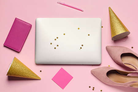 Laptop with high-heeled shoes and party hats on color background