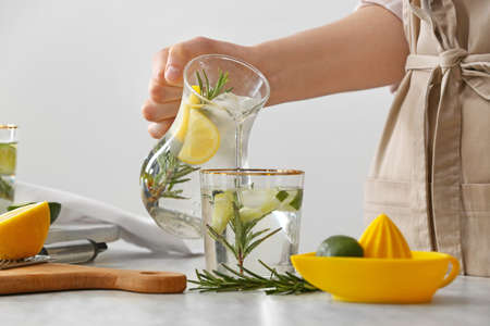 Woman pouring lime cocktail with rosemary into glass on table in kitchenの写真素材