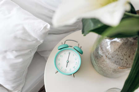 Stylish alarm clock and vase with flowers on table in bedroom