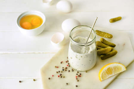 Glass jar with tasty tartare sauce and ingredients on light wooden background
