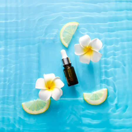 Cosmetic dropper bottle with flowers and lemon in water on color background