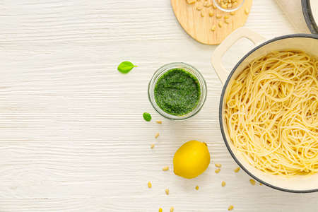 Jar of tasty pesto sauce, cooking pot of pasta and ingredients on light backgroundの写真素材